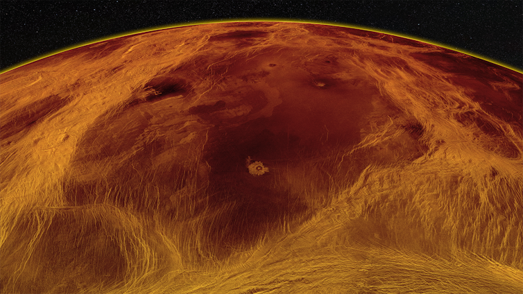 A red–hued image of the surface of Venus showing a large darker piece of the surface surrounded by lighter colors against the backdrop of space.