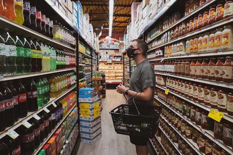 A man in a mask surveys grocery store shelves while carrying a basket.