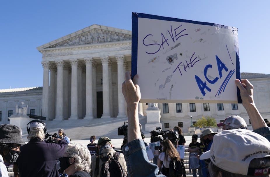 In the foreground is a handwritten sign, reading "Save the ACA." The Supreme Court building is in the background.