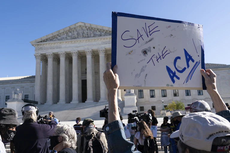 In the foreground is a handwritten sign, reading "Save the ACA." The Supreme Court building is in the background.