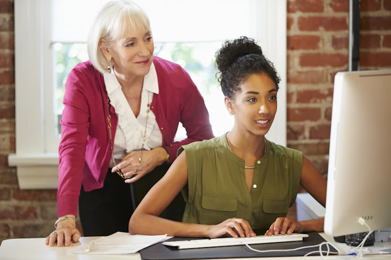 Older woman mentoring a younger women looks over her shoulder as she works at a PC