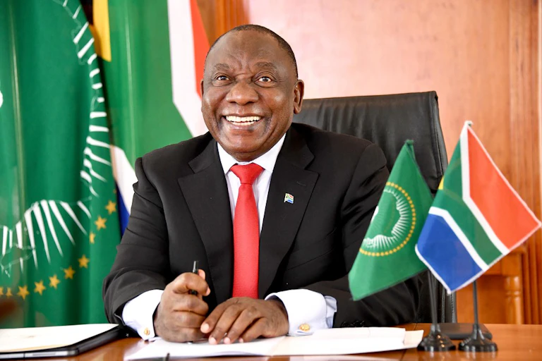 Smiling man wearing suit and tie, seated at desk; flags on the desk and in the background