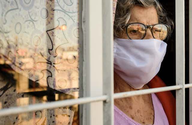 Una mujer mayor pensativa con mascarilla y gafas se asoma a una ventana.