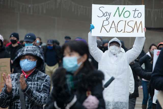 A person wearing a mask holds a sign reading Say No to Racism at a protest
