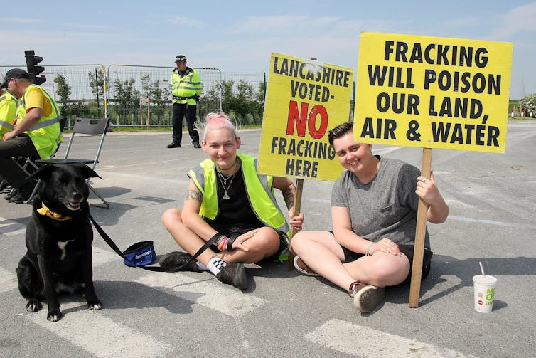 Two girls sit on the tarmac with yellow anti-fracking placards and a dog on a leash