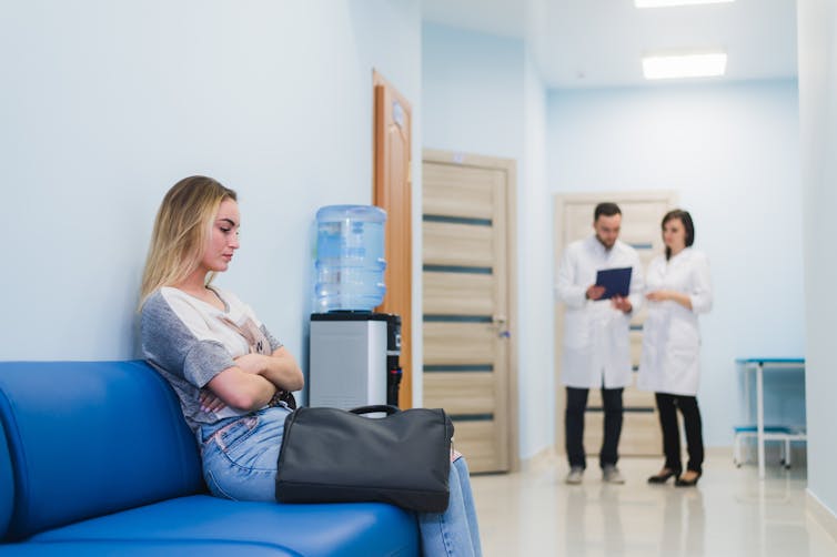 Young woman waiting alone at doctor's office, while two doctors consult in the background.