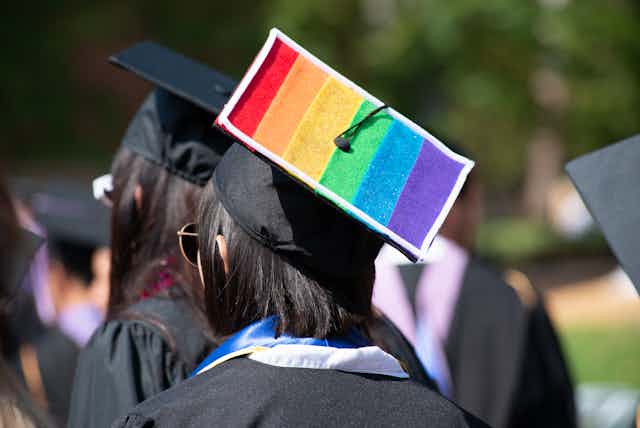 A graduating student wears a black gown and cap with the rainbow flag on it.