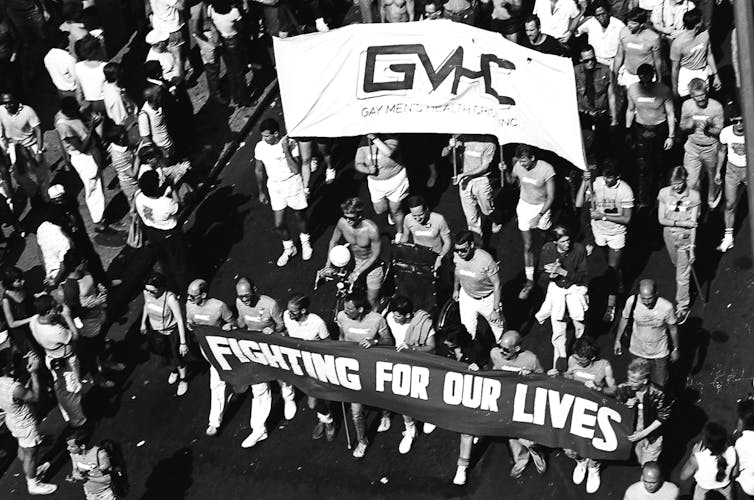 Men march shirtless holding a 'GMHC' lbanner and another that reads 'Fighting for Our Lives,'