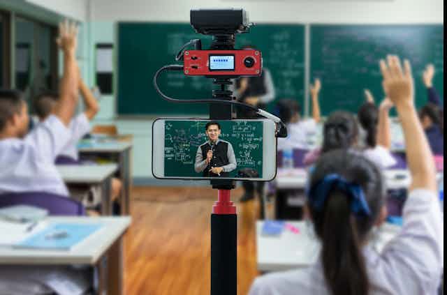 Un professeur qui donne un cours dans une classe est filmé en même temps par un téléphone.