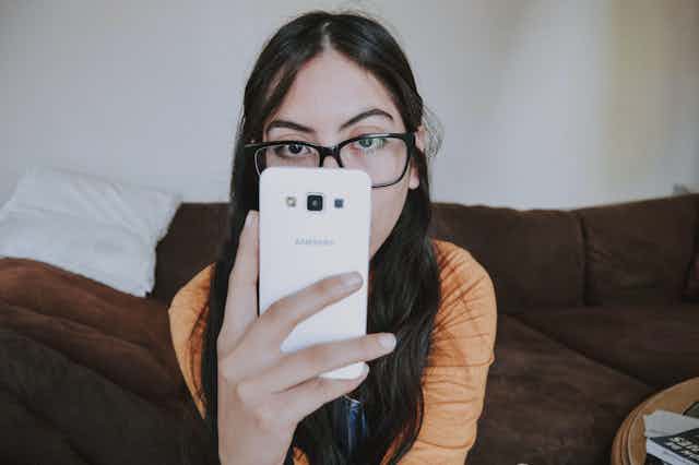 A young woman with long dark hair and glasses peers out at the viewer, a smart phone in her hand covers her nose and mouth