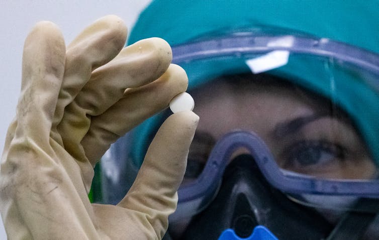Person in PPE holds a white pill up in front of their eye.