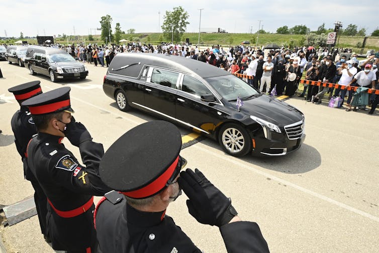 Police officers salute as a hearse drives by, muslim community members and supporters stand in the background