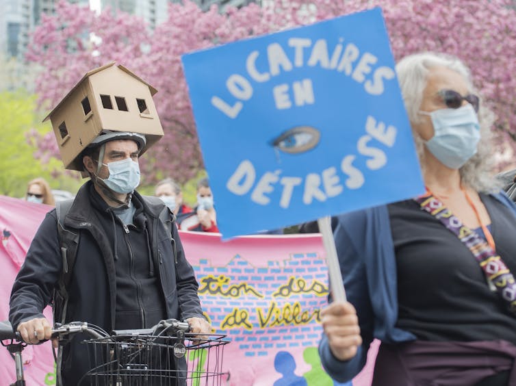 A man wears a hat in the shape of a house and a women holds a sign at a march