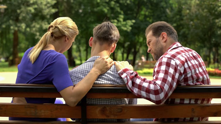 Parents sit on bench with son in the middle, hands are on his shoulder