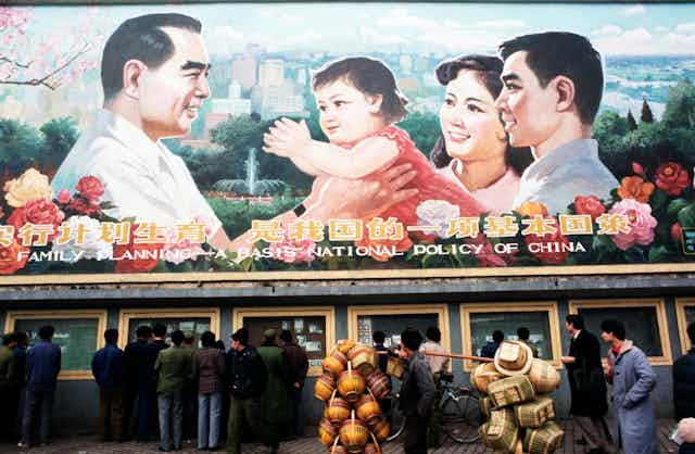Pedestrians and a man carrying baskets pass by a huge billboard extolling the virtues of China's 'One Child Family' policy.