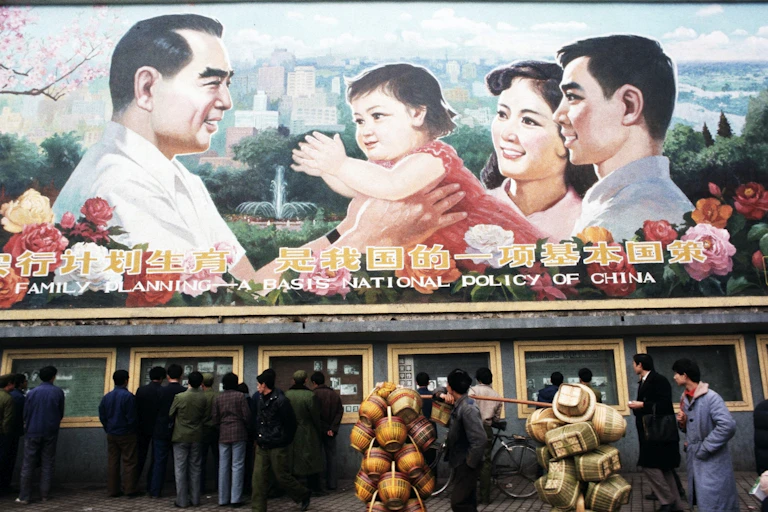 Pedestrians and a man carrying baskets pass by a huge billboard extolling the virtues of China's 'One Child Family' policy.