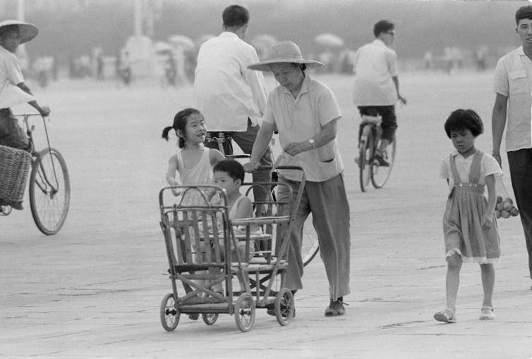 A mother wearing a straw hat pushes a buggy with a child sitting it. There are two other children walking near the buggy.