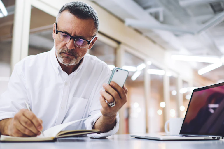 middle-aged man with phone and laptop making notes