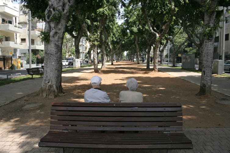 Elderly couple sit on a bench in shade under street trees