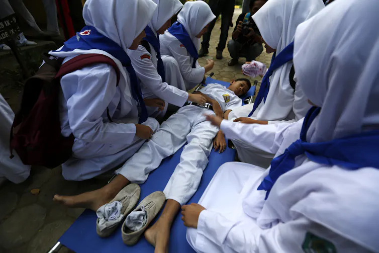 Schoolchildren in white outfits cluster around a young boy pretending to be injured.