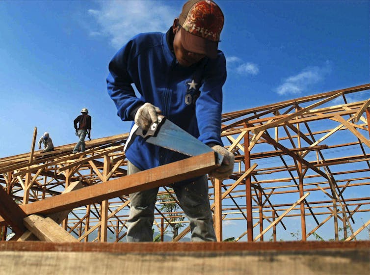 A man in a red cap saws wood, the frame of a building in the background.