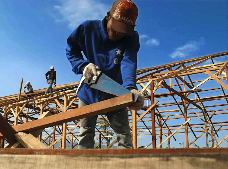 A man in a red cap saws wood, the frame of a building in the background.