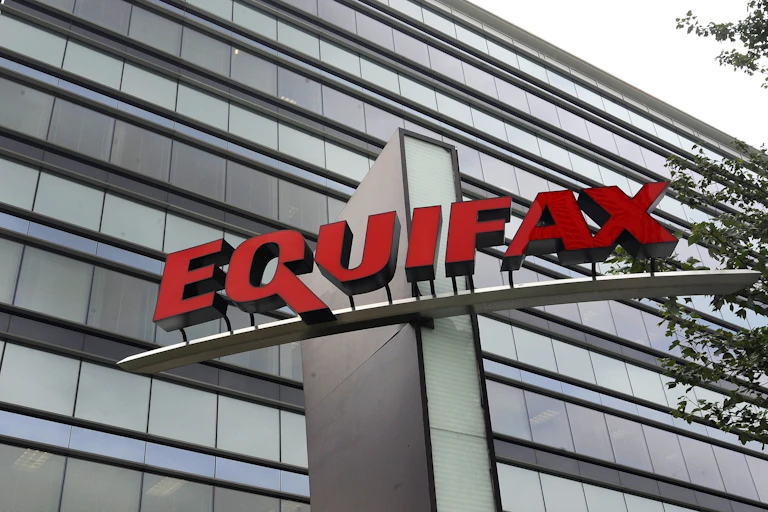 The glass façade of a large office building with a large sign mounted on a structure in front of the building bearing the name Equifax