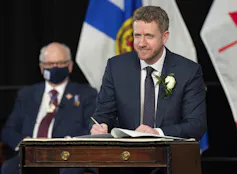 A man standing at a podium in front of the Nova Scotia flag