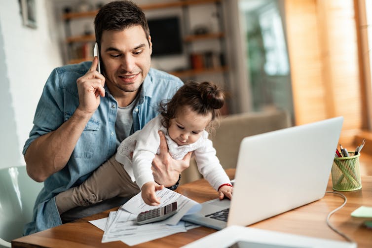 Father on the phone in front of his laptop while holding kid who is trying to touch everything on the desk.