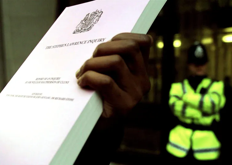 Hand holding up large document with the words STEPHEN LAWRENCE INQUIRY on the front while a police officer stands in the background