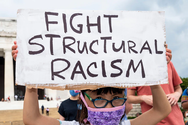 Little girl wearing glasses and a face mask holds up sign that says 'fight structural racism'