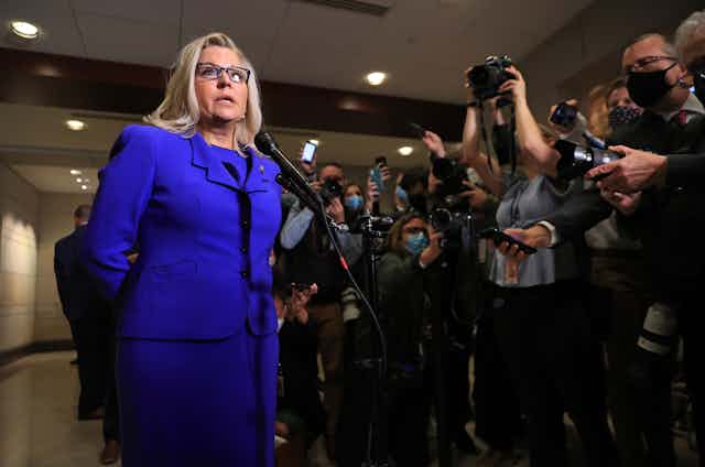 Liz Cheney, a blond woman with glasses and wearing a blue suit, talking to a large crowd of reporters