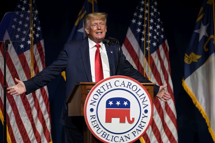 President Donald Trump in a blue suit and red tie, standing in front of two American flags and behind a lectern with the symbol of the North Carolina Republican Party.