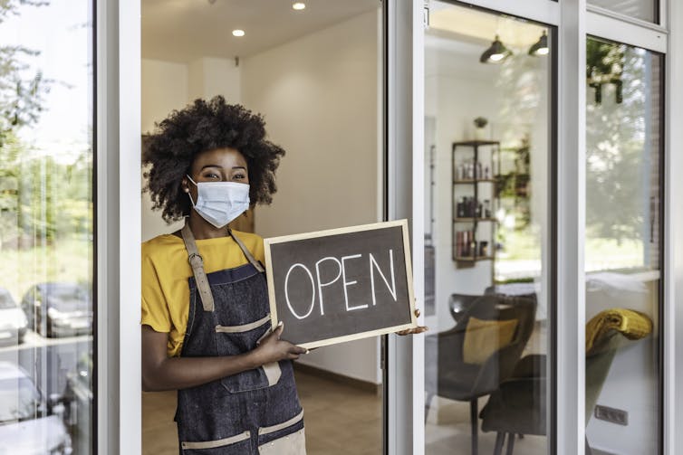 Una mujer afroamericana con máscara protectora quirúrgica con cartel abierto frente a su pequeña tienda.
