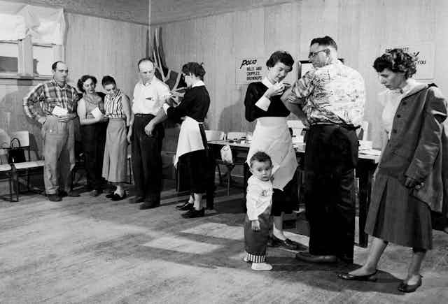 Black and white photo, nurse giving polio vaccine to man as child tugs on her apron
