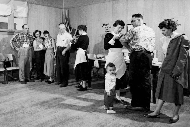 Black and white photo, nurse giving polio vaccine to man as child tugs on her apron