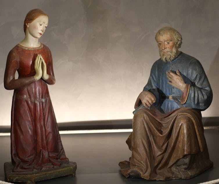 A woman in a simple red dress with hands folded in prayer next to a kneeling man.