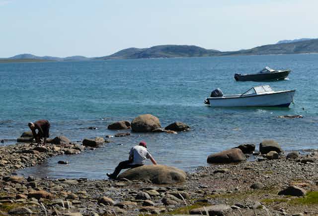 A rocky shore with two people in the foreground and two boats in the water in the background