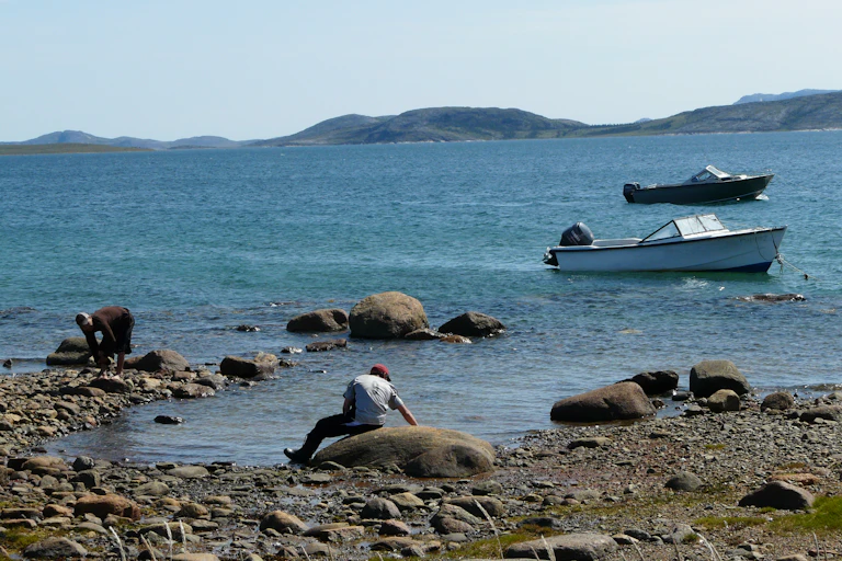 A rocky shore with two people in the foreground and two boats in the water in the background
