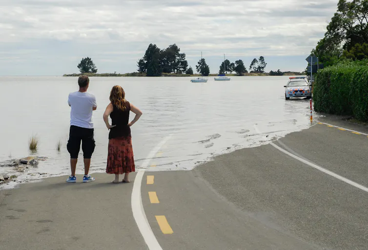 Two people look on as a motorway is engulfed by ocean water.