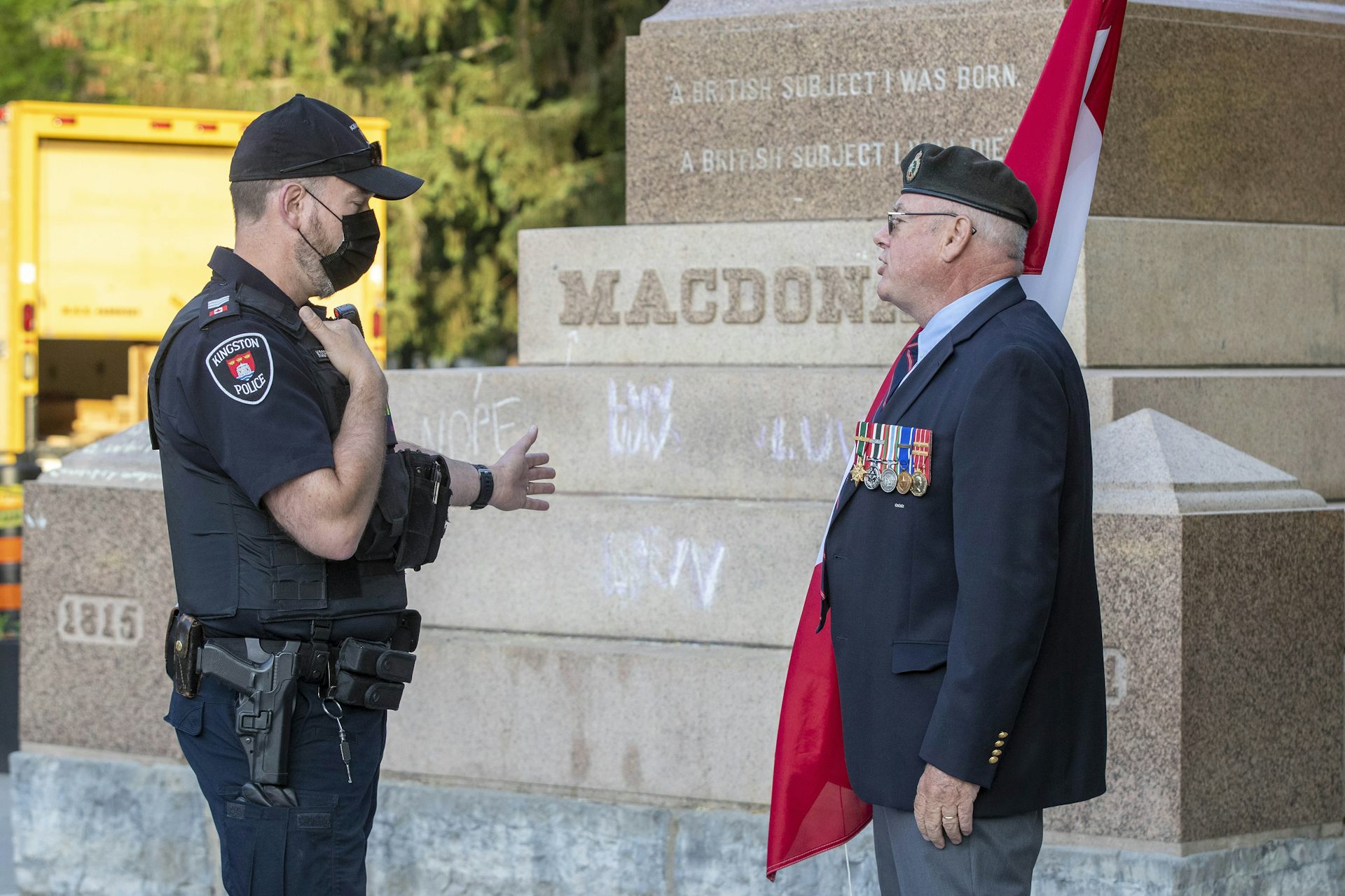 A police officer gestures with to a statue while talking with a veteran holding a Canadian flag.