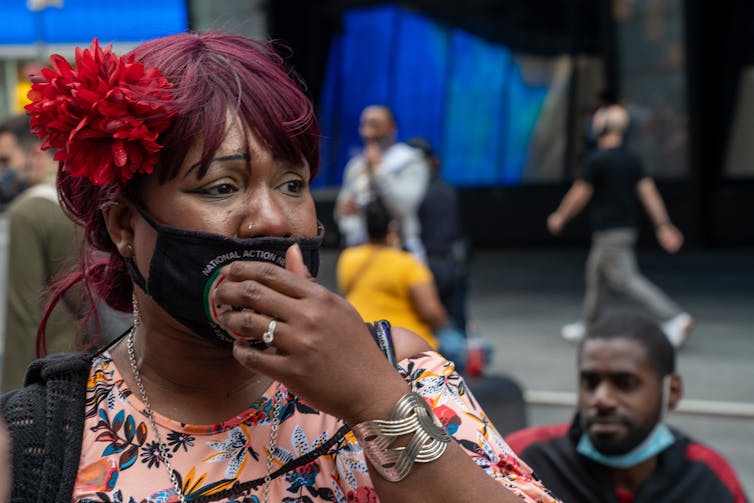 Black woman in a face masks cries on a city street, with a hand over her mouth