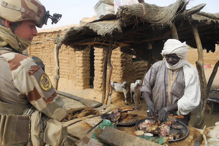 A soldier in full combat gear stands in front of a man arranging meat on a table