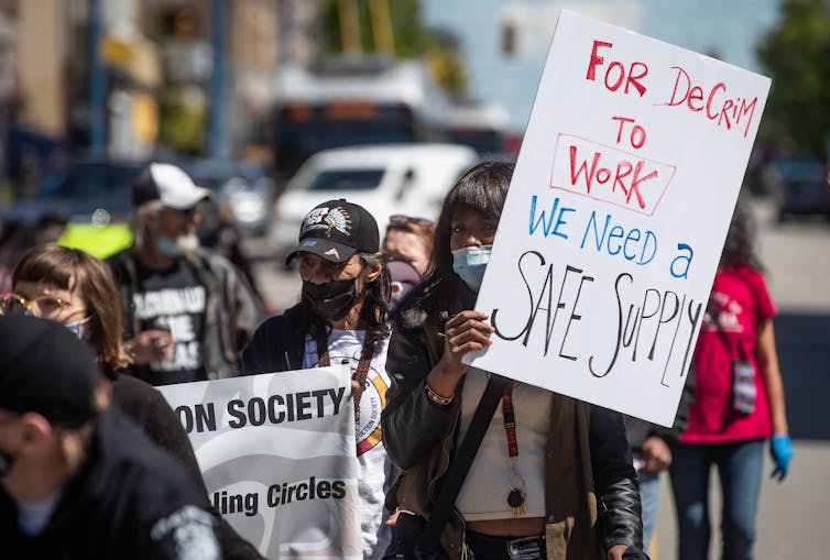 A woman holds a sign during a protest reading FOR DECRIM TO WORK WE NEED A SAFE SUPPLY