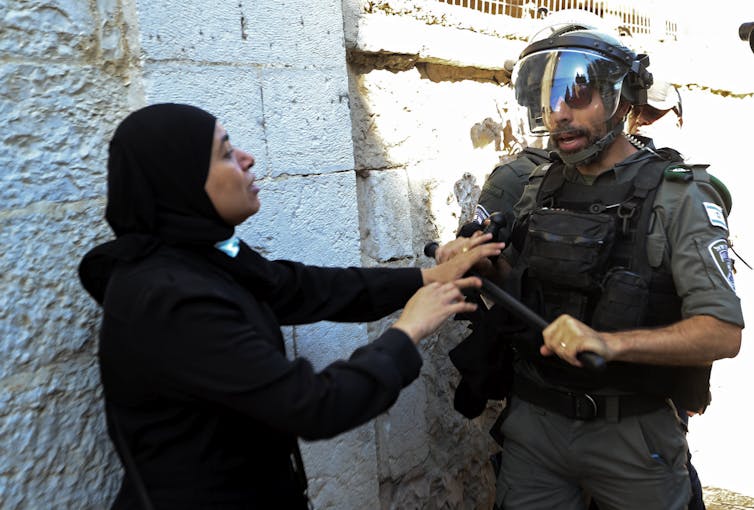 A woman raises her hands as a heavily armoured police officer confronts her.