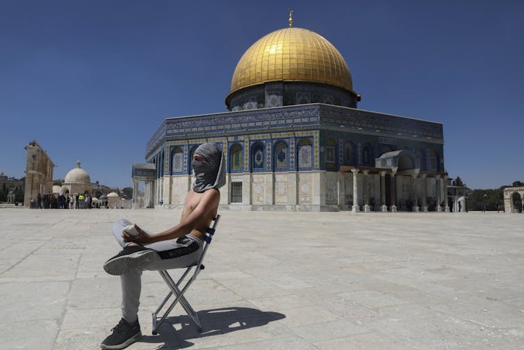 A man in a chair holding a rock sits in the foreground with the Dome of the Rock Mosque in the background.