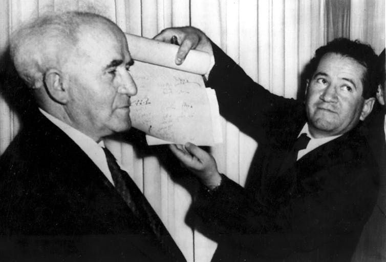 A man unrolls a scroll as David Ben-Gurion looks on.
