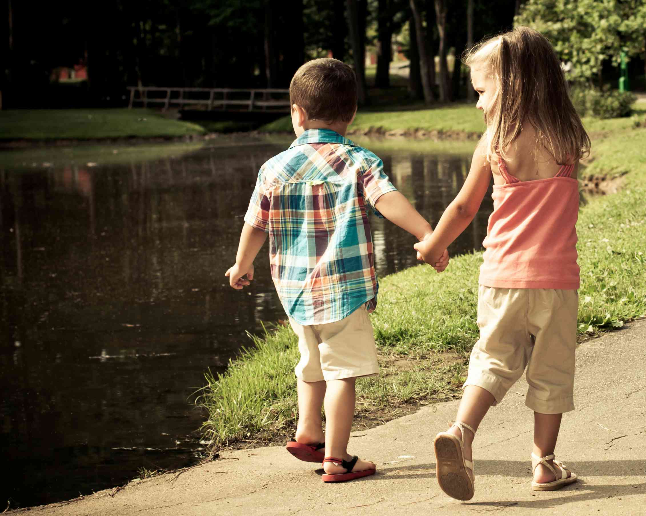 Brother and sister holding hands walking past a lake.