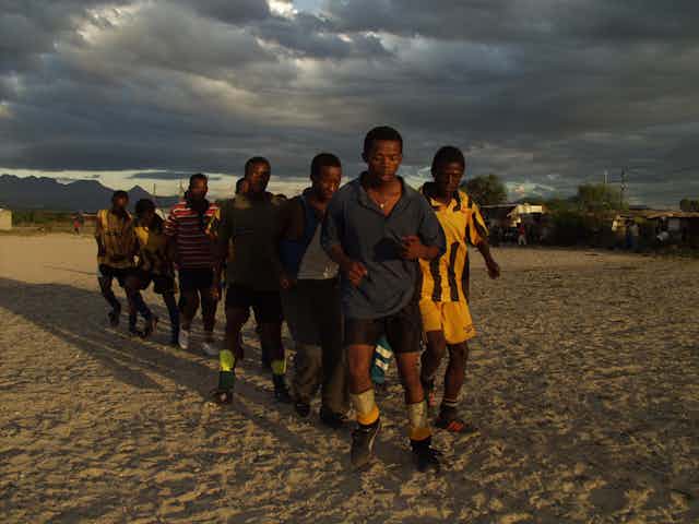A pack of young men in football clothes form a triangle as they jog towards camera, late afternoon skies.