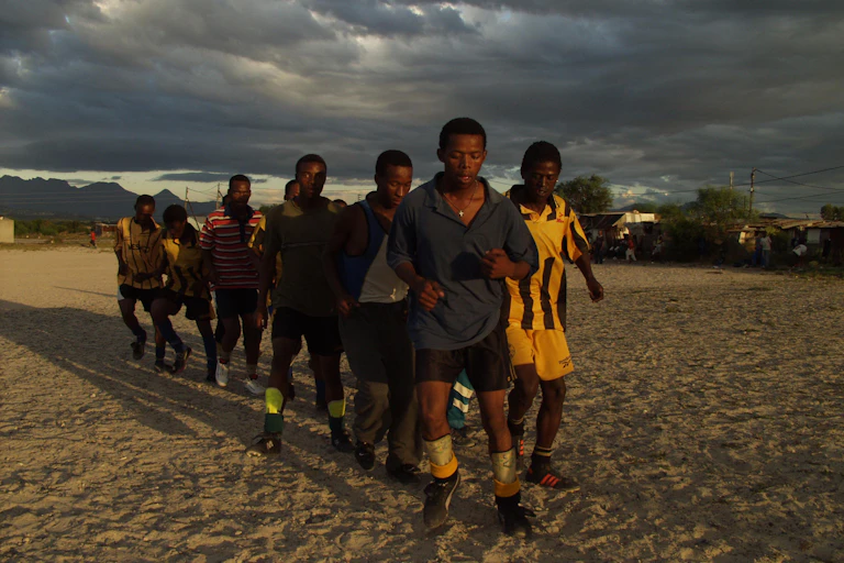A pack of young men in football clothes form a triangle as they jog towards camera, late afternoon skies.